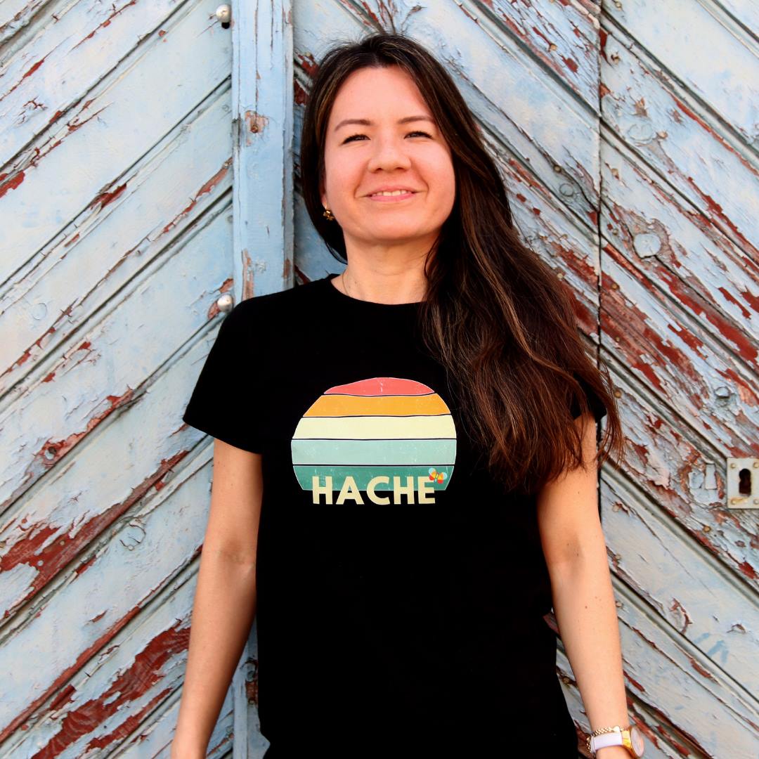 Una chica posa con una camiseta negra con un semicírculo partido en filas de diferentes colores y con la palabra hache en color crema en la fila inferior.