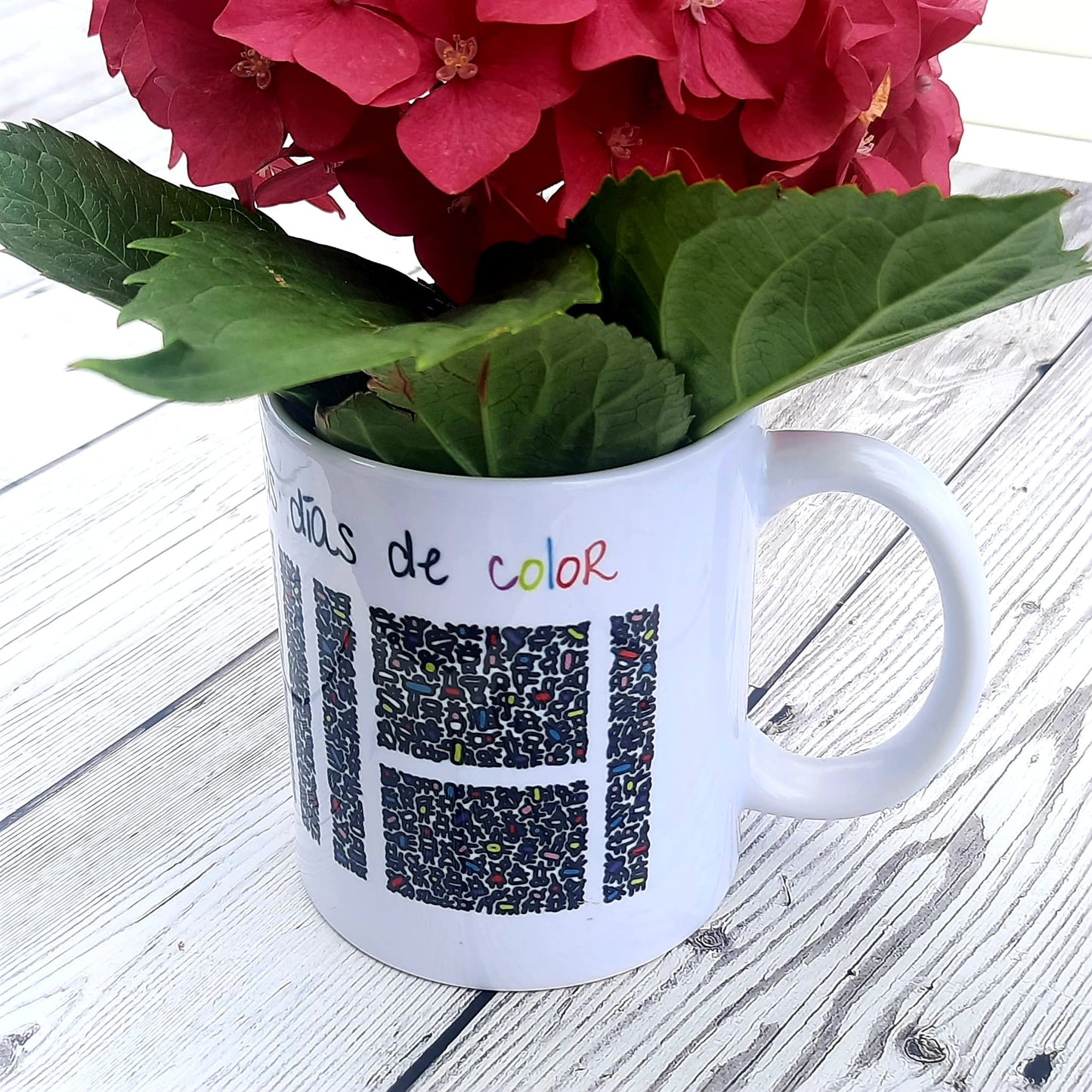 Taza blanca con figuras abstractas de colores que por su disposición espacial forman una Hache blanca en medio. Encima, la frase Gracias por llenar mis días de color.