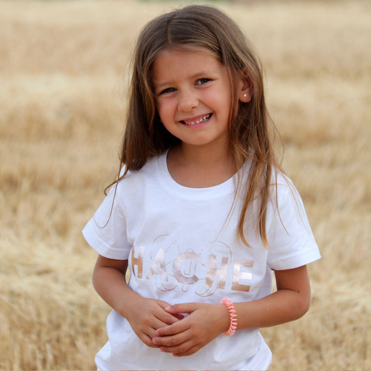 Niña con camiseta blanca con palabra HACHE escrita en rosa plateado y silueta de mariposa detrás.