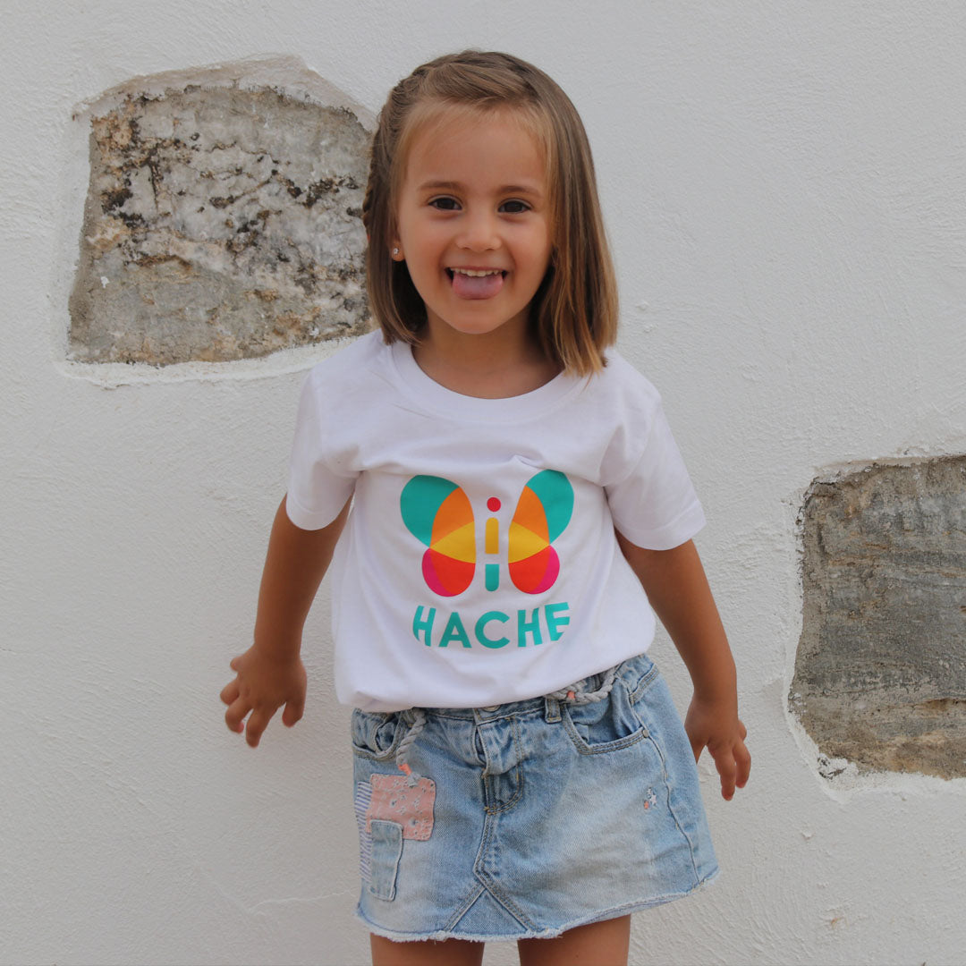 Niña con camiseta blanca. Mariposa HACHE central multicolor con palabra HACHE escrita en azul debajo