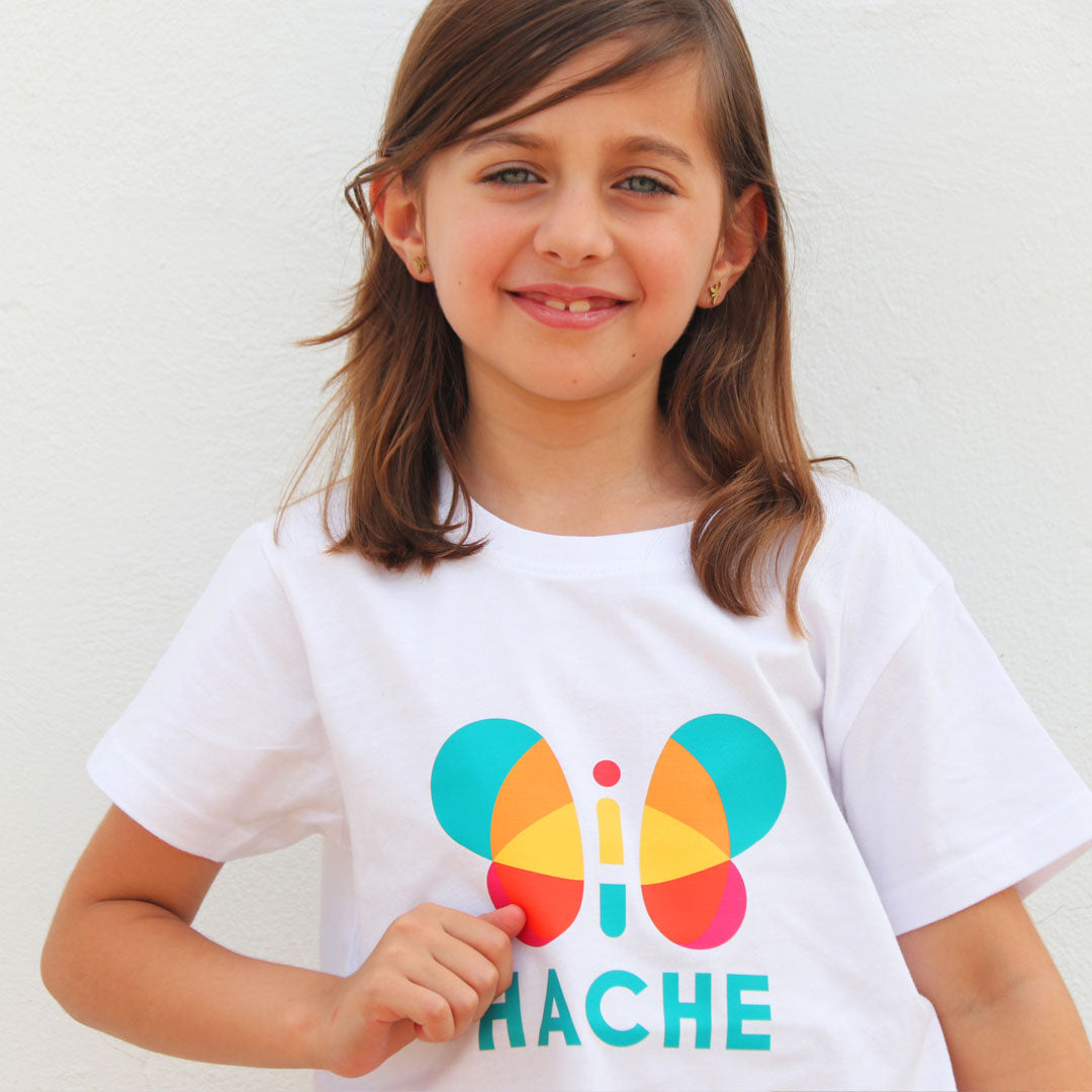 Niña con camiseta blanca. Mariposa HACHE central multicolor con palabra HACHE escrita en azul debajo