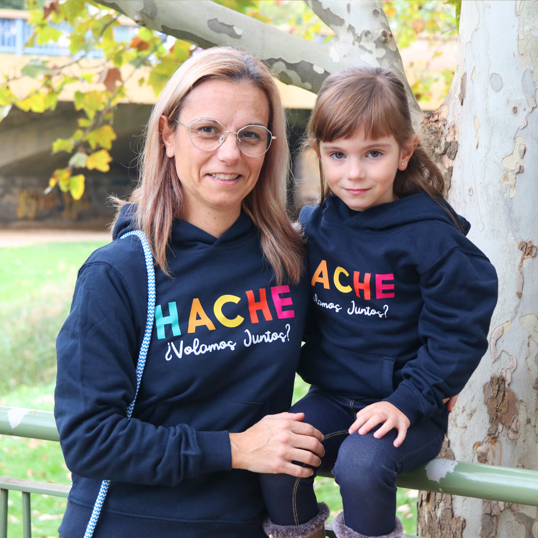 Niña y madre lucen sudadera azul oscura con Hache multicolor y frase Volamos Juntos en blanco debajo