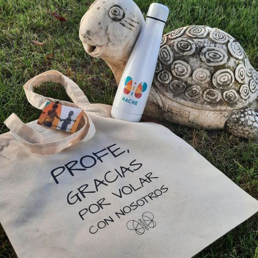 Botella blanca con logo HACHE a color y bolsa de tela con texto "profe, gracias por volar con nosotros".