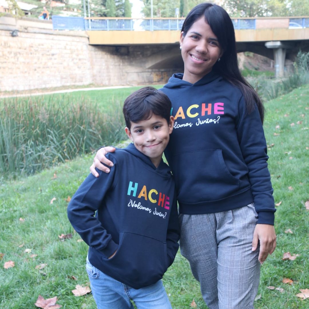 Niño y madre lucen sudadera azul oscura con Hache multicolor y frase Volamos Juntos en blanco debajo