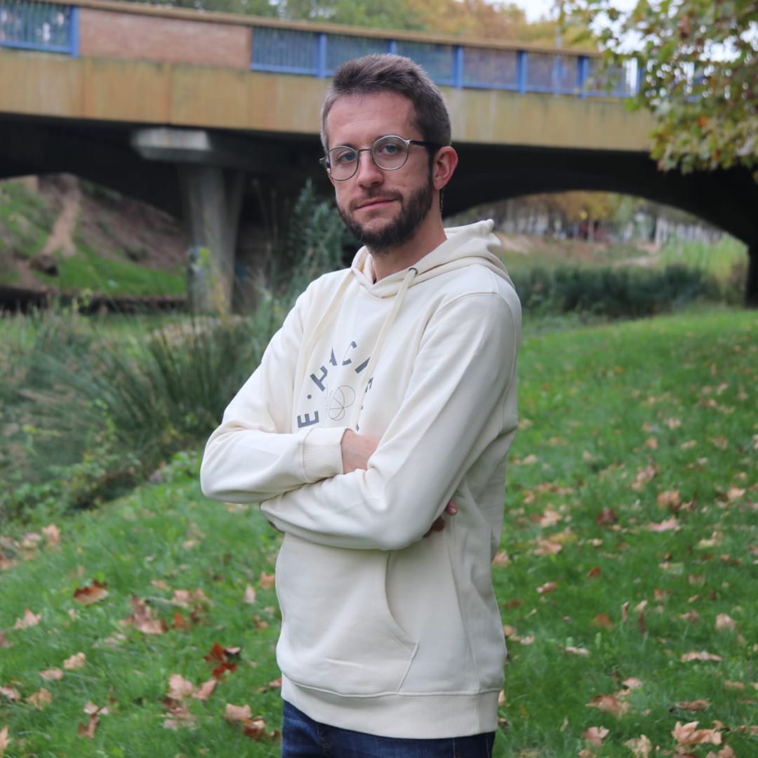 Chico posa con los brazos cruzados con sudadera crema y círculo hecho con las letras de la palabra Hache y una mariposa en silueta en el centro.