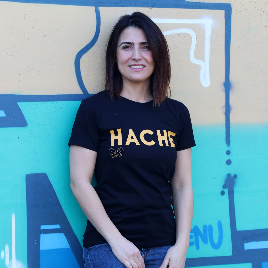chica con camiseta color negra de manga corta con palabra HACHE en mostaza y tamaño grande en el pecho y silueta de mariposa en color mostaza debajo de la letra H
