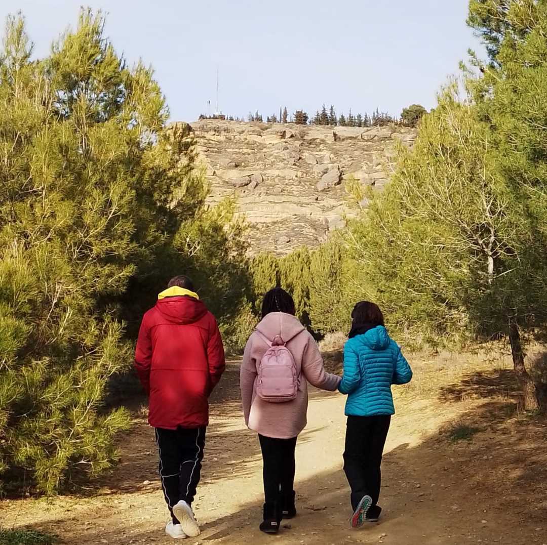 Un chico y dos chicas pasean por un camino de tierra. A los lados hay árboles. La foto está hecha de espaldas