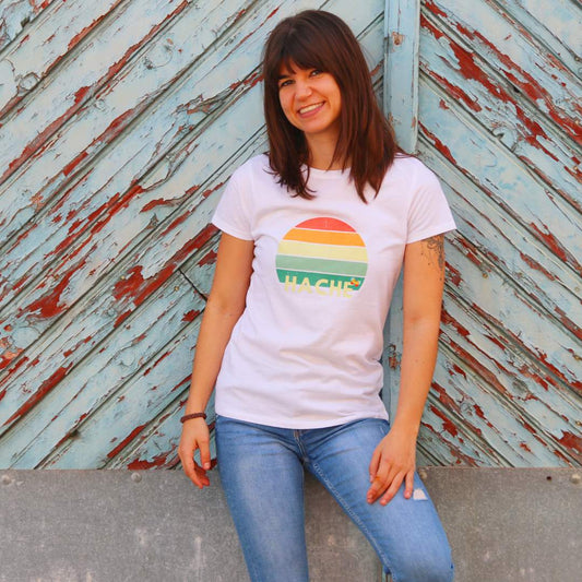 Una chica posa con una camiseta blanca con un semicírculo partido en filas de diferentes colores y con la palabra hache en color crema en la fila inferior.