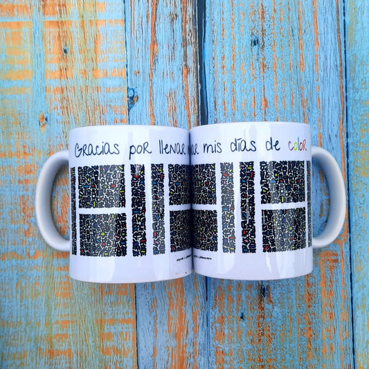 Taza blanca con figuras abstractas de colores que por su disposición espacial forman una Hache blanca en medio. Encima, la frase Gracias por llenar mis días de color.