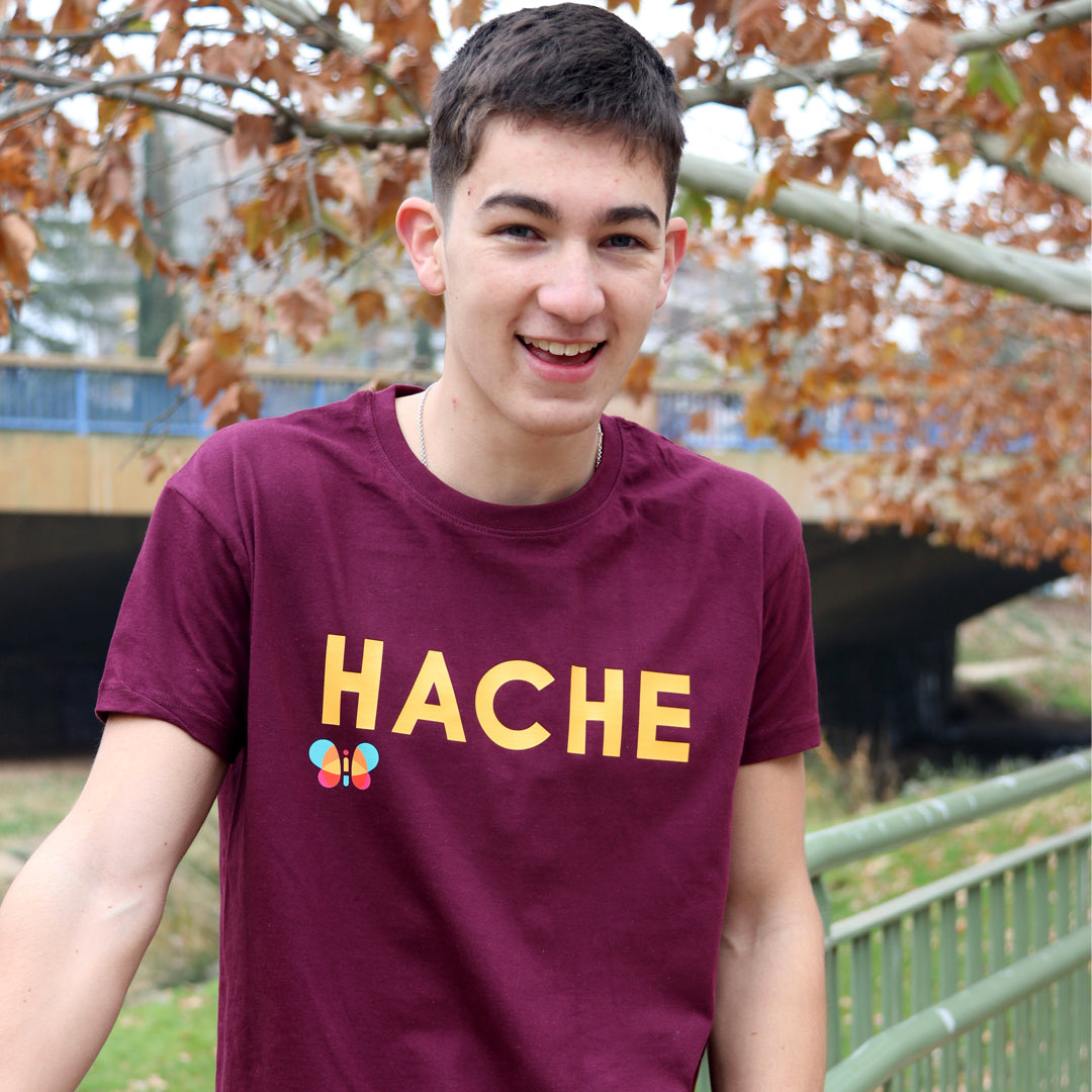 chico con camiseta color burdeos de manga corta con palabra HACHE en mostaza y tamaño grande en el pecho y mariposa a color debajo de la letra H