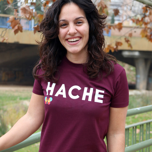 chica con camiseta color burdeos de manga corta con palabra HACHE en blanco y tamaño grande en el pecho y mariposa a color debajo de la letra H