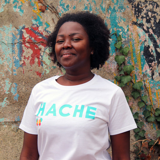 chica con camiseta color blanca de manga corta con palabra HACHE en azul cielo y tamaño grande en el pecho y mariposa a color debajo de la letra H