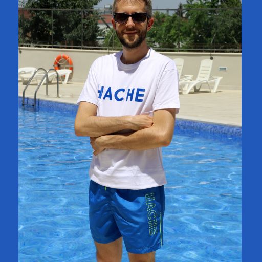 Modelo con camiseta blanca y palabra HACHE escrita en azul en el pecho y bañador azul eléctrico con palabra HACHE escrita en verde lima en un lateral en sentido vertical