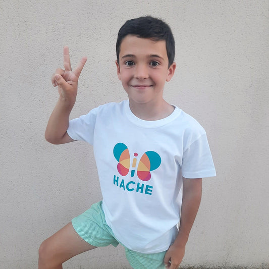 Niño con camiseta blanca. Mariposa HACHE central multicolor con palabra HACHE escrita en azul debajo