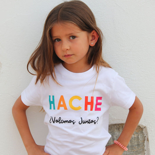 Niña con camiseta blanca. Palabra HACHE escrita en el pecho con un color de cada letra (azul, naranja, amarillo, rojo y rosa) y la pregunta ¿Volamos juntos? escrita en negro debajo