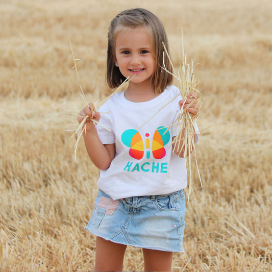 Niña con camiseta blanca. Mariposa HACHE central multicolor con palabra HACHE escrita en azul debajo