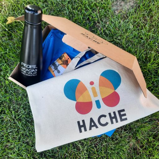 Bolsa de tela con logo HACHE a color y botella negra con texto "profe, gracias por volar con nosotros". Todo colocado dentro de una caja de cartón