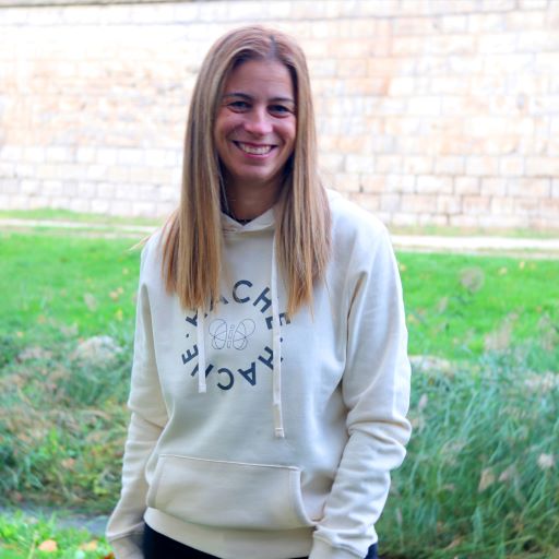 Chica posa sonriendo con sudadera crema y círculo hecho con las letras de la palabra Hache y una mariposa en silueta en el centro.