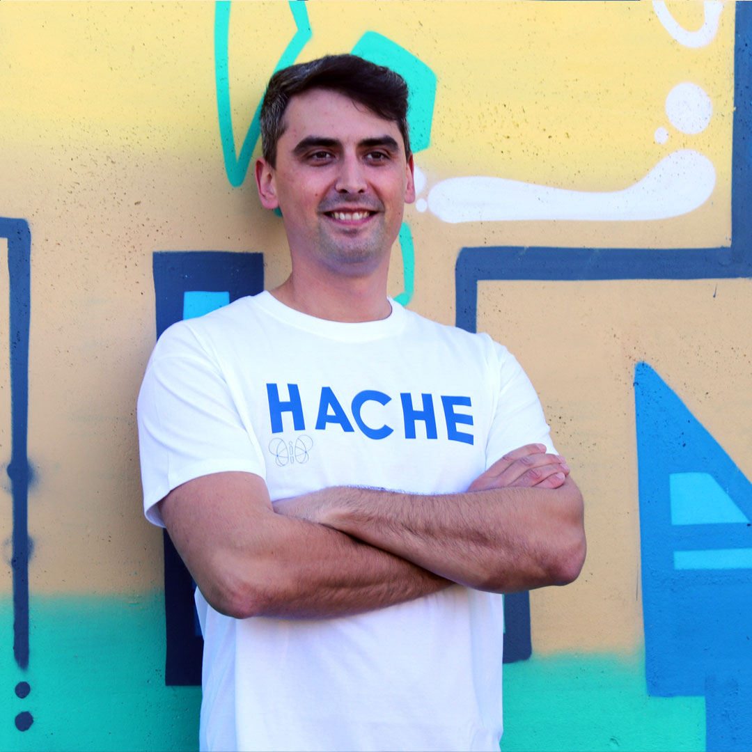 chico con camiseta color blanca de manga corta con palabra HACHE en azul y tamaño grande en el pecho y mariposa en silueta azul debajo de la letra H